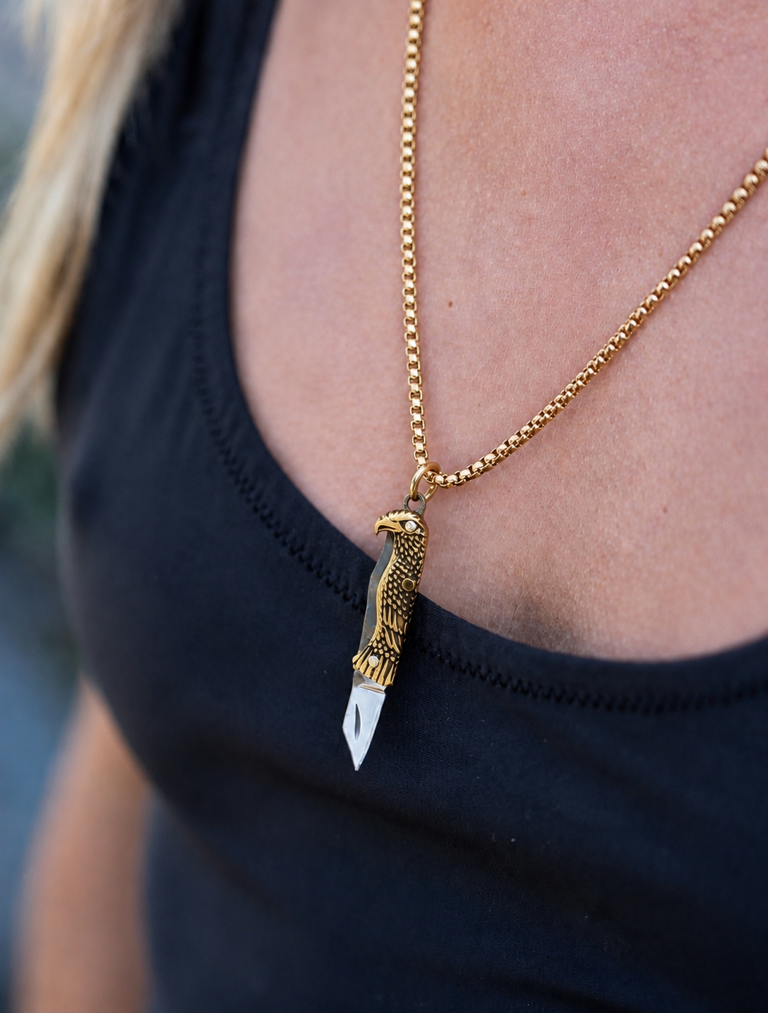 A close-up of a gold chain necklace with a detailed eagle pendant holding a small silver knife blade, worn by a person in a black top.