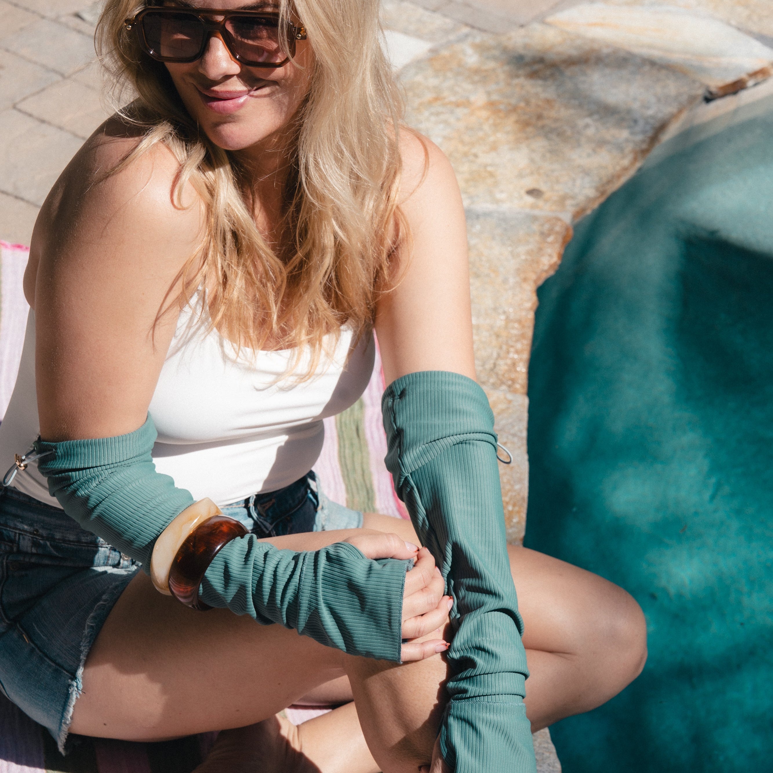 Woman sitting outdoors wearing teal arm sleeves and sunglasses.