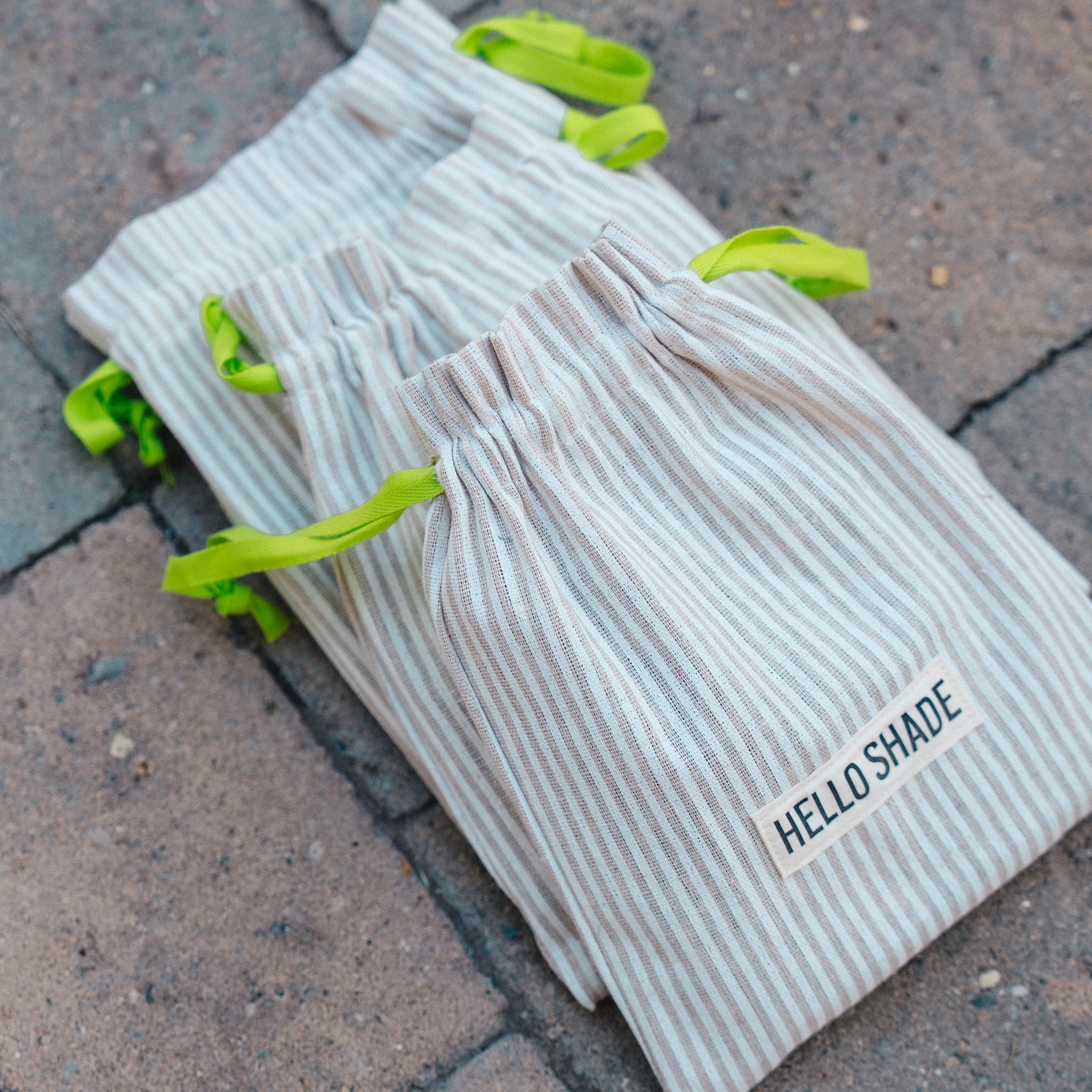 Stack of linen pouches with neon green straps on a brick surface, featuring 'HELLO SHADE' branding.