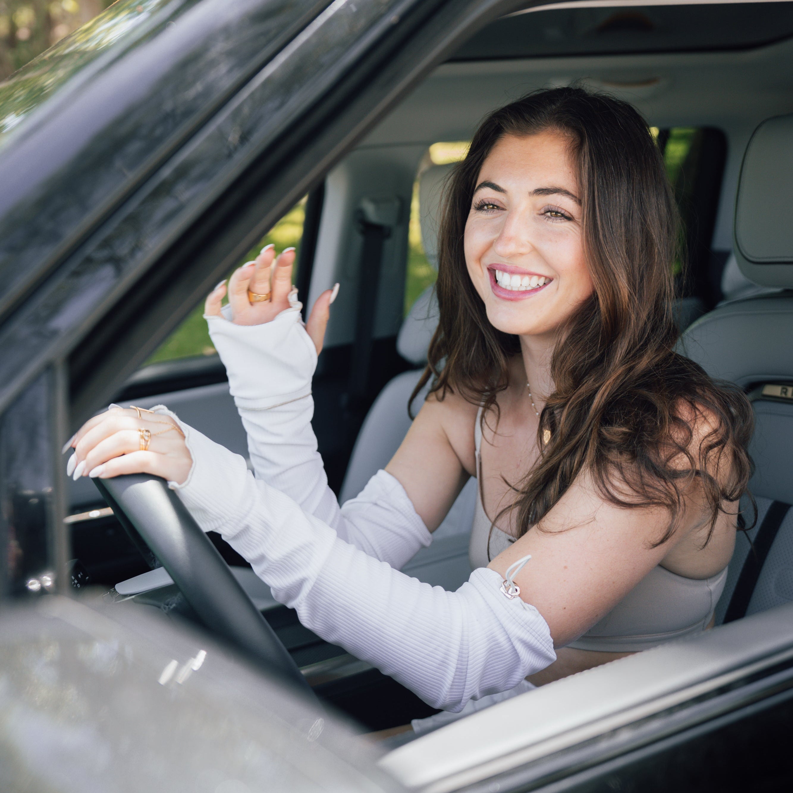 A smiling woman with long brown hair sits in the driver's seat of a car, wearing a white sleeveless top and white arm sleeves. The car interior is gray, and trees are visible through the background window.