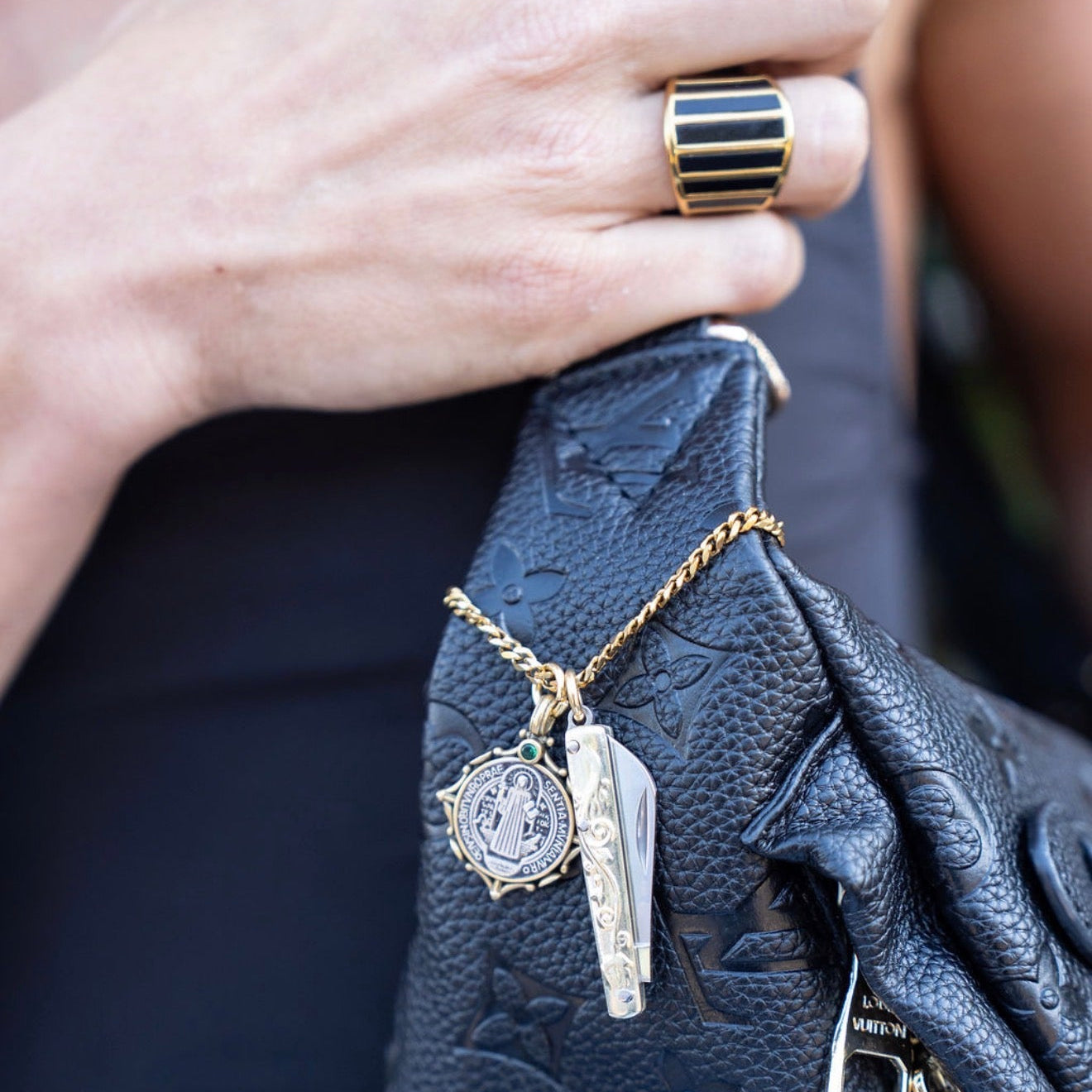 Close-up of a hand holding a black textured bag with a gold necklace featuring two pendants.