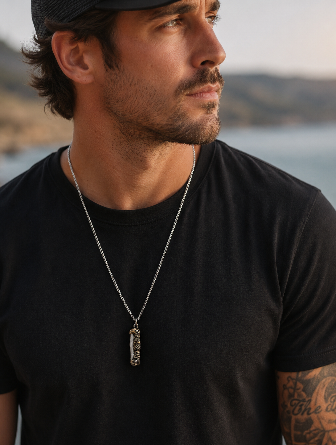 A man wearing a silver-toned chain necklace with a eagle knife pendant featuring a textured, metallic interior, photographed outdoors against a blurred background of water and hills.